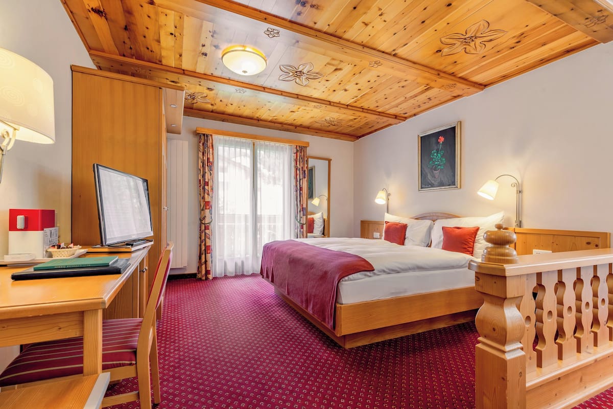 Una habitación de hotel con techo de madera, cama con manta roja, escritorio con computadora y ventana con cortinas.