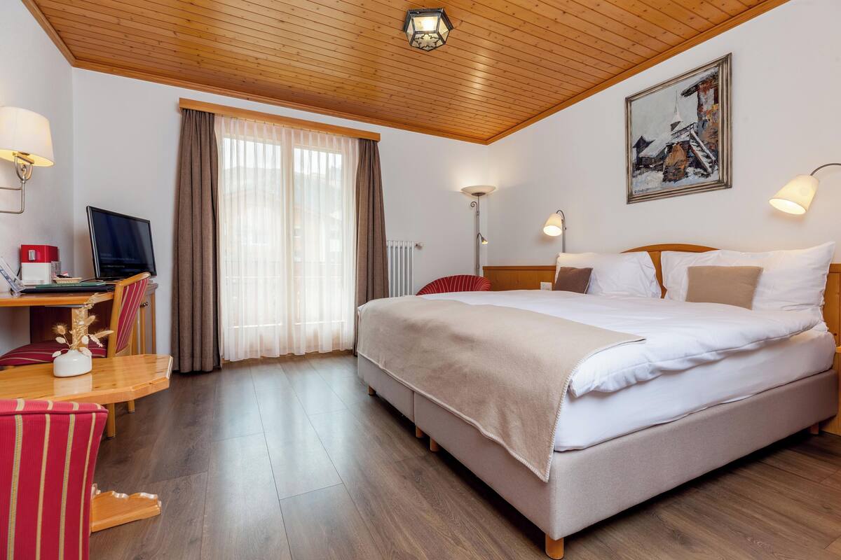 Una habitación de hotel con techo de madera, cama con ropa blanca, escritorio con ordenador, una silla y ventana con cortinas.