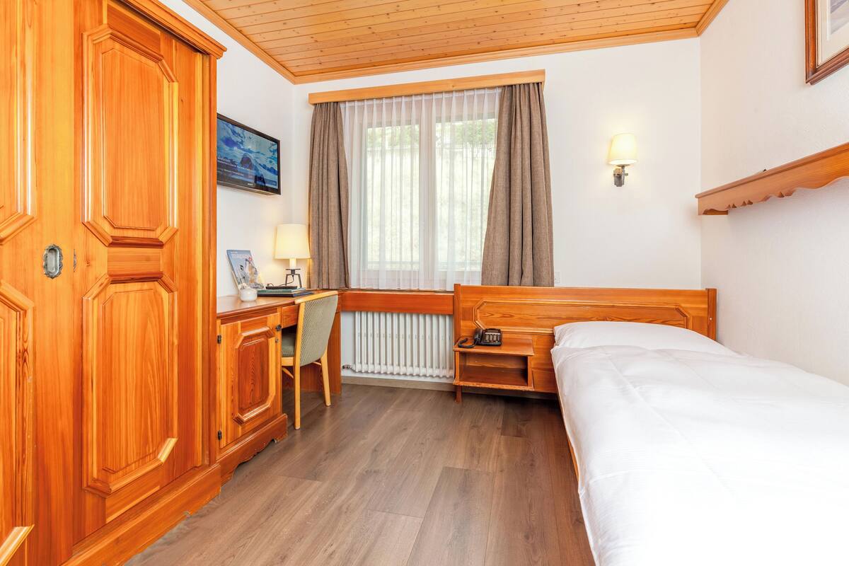 Habitación de hotel con un armario de madera, un escritorio con lámpara, una cama y una ventana con cortinas.