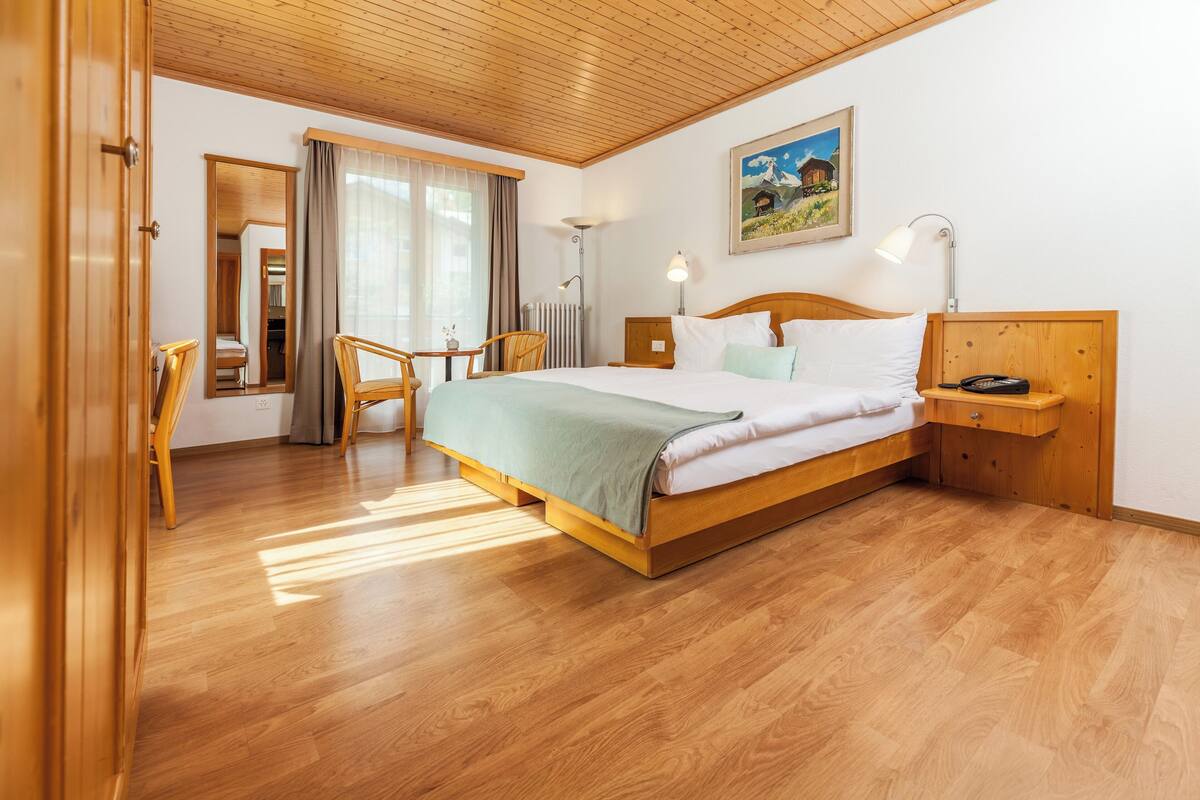 Habitación de hotel con una cama de madera, un escritorio, una silla y un amplio ventanal con cortinas.
