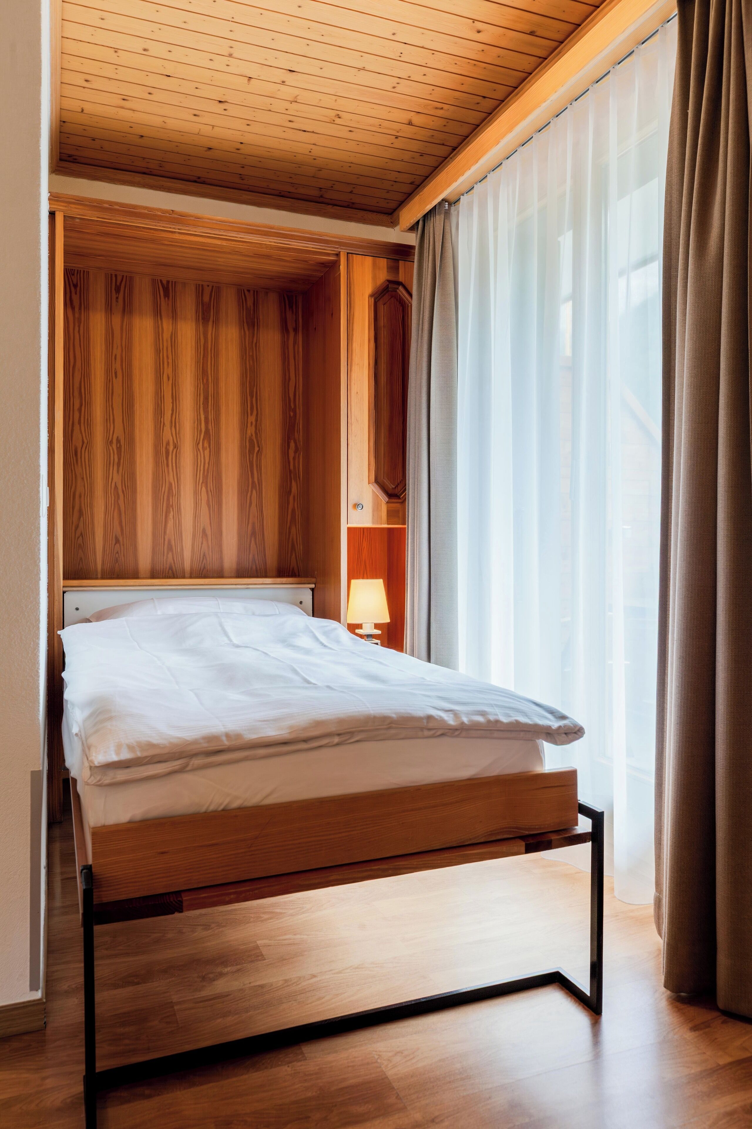 A single bed with a wooden frame and headboard, a bedside table with a lamp, and a window with sheer curtains.