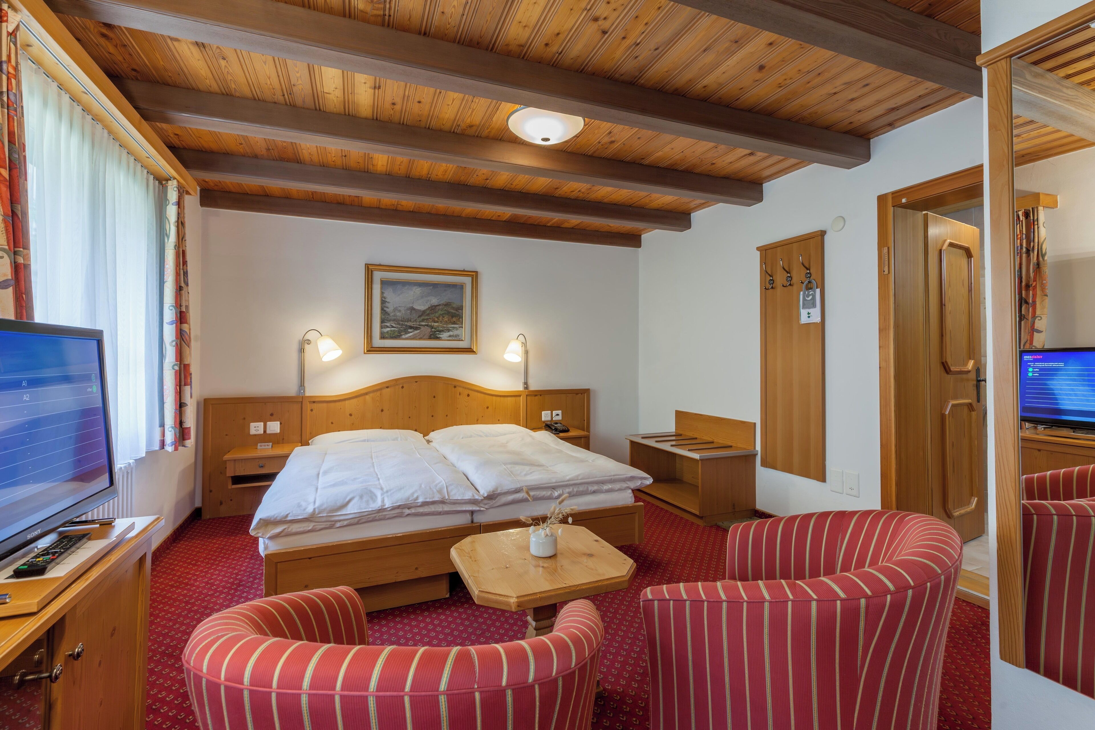 Ett hotellrum med ett träpanelat tak, en säng med vita sängkläder, en tv, ett skrivbord med en dator och randiga fåtöljer.