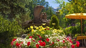 A garden with a large planter of red and white flowers, a stone sculpture, and a yellow umbrella. - Alpenblick (Zermatt)