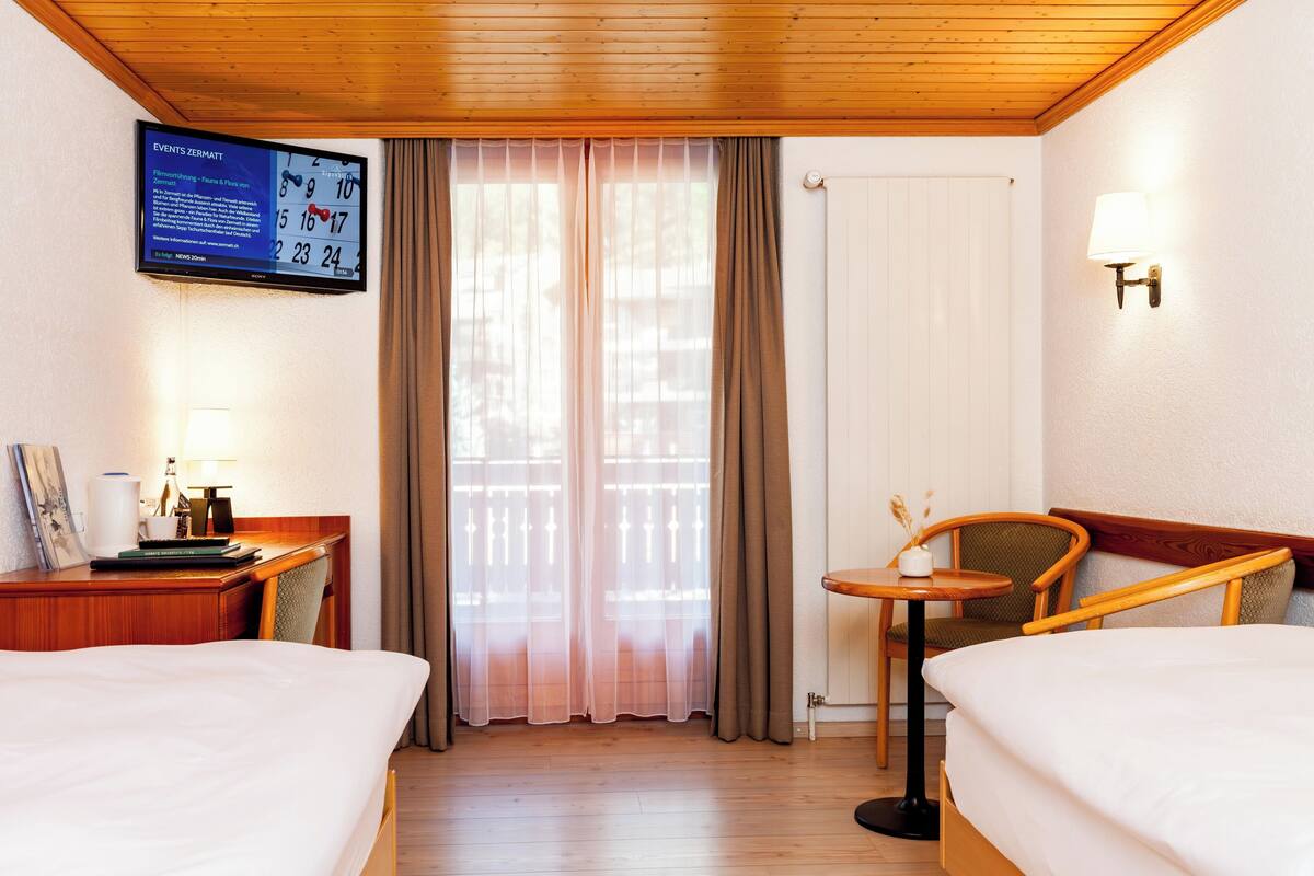 Habitación de hotel con dos camas, un escritorio con lámpara, un televisor mostrando información de eventos y vistas al exterior a través de una ventana con cortinas.