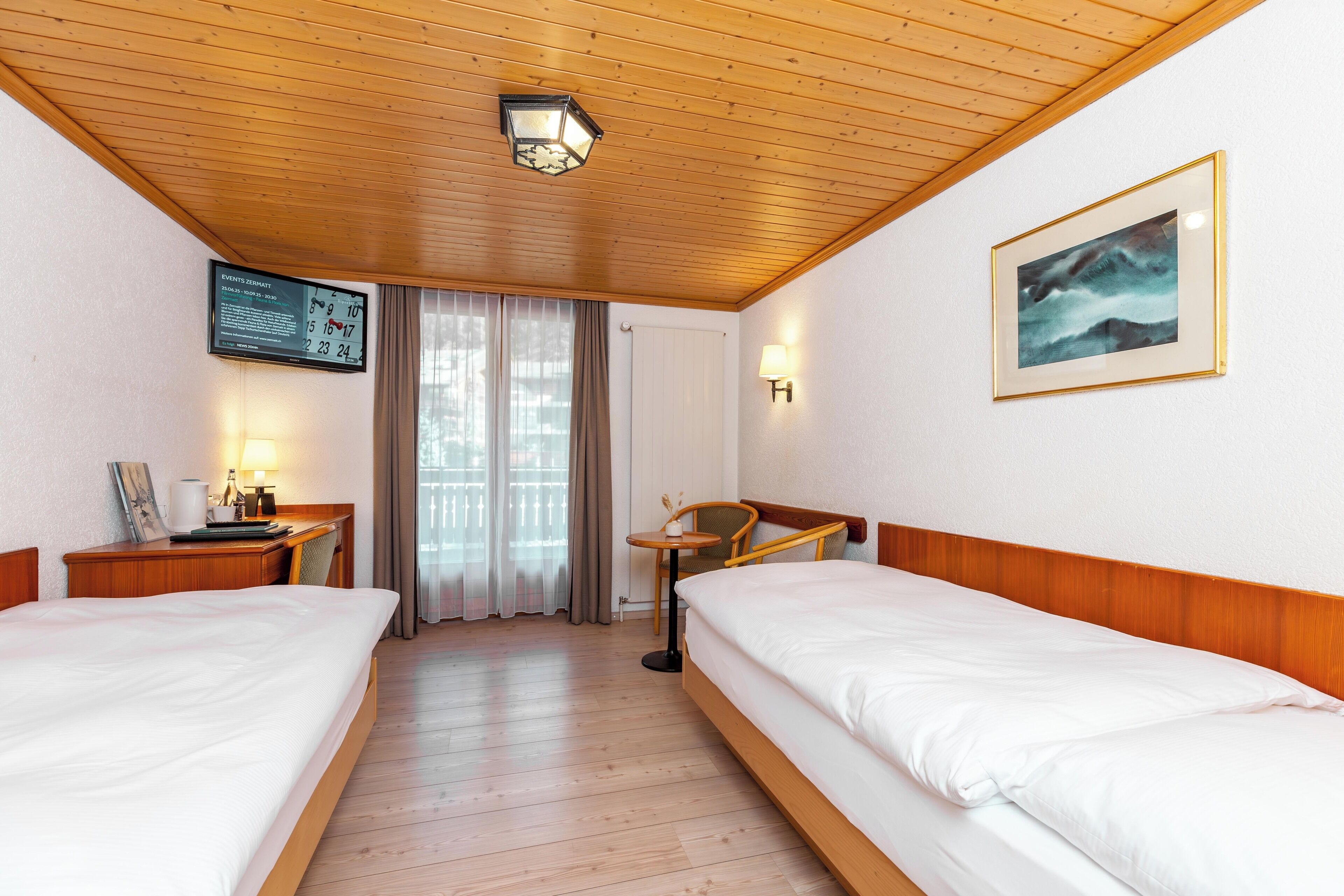 Quarto de hotel com duas camas, uma escrivaninha com luminária, uma mesinha e uma televisão fixada na parede, além de um quadro emoldurado.