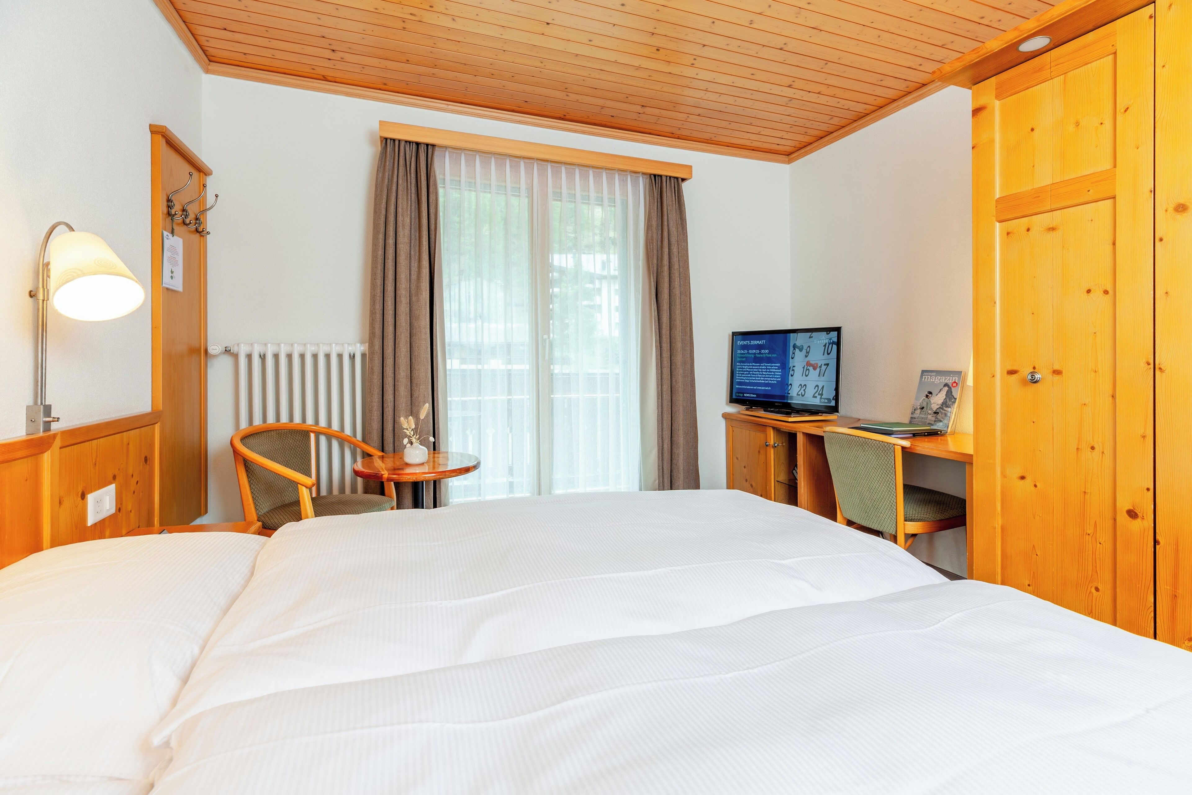 A hotel room with a large bed, a desk with a computer, a chair, a lamp, and a window with curtains.
