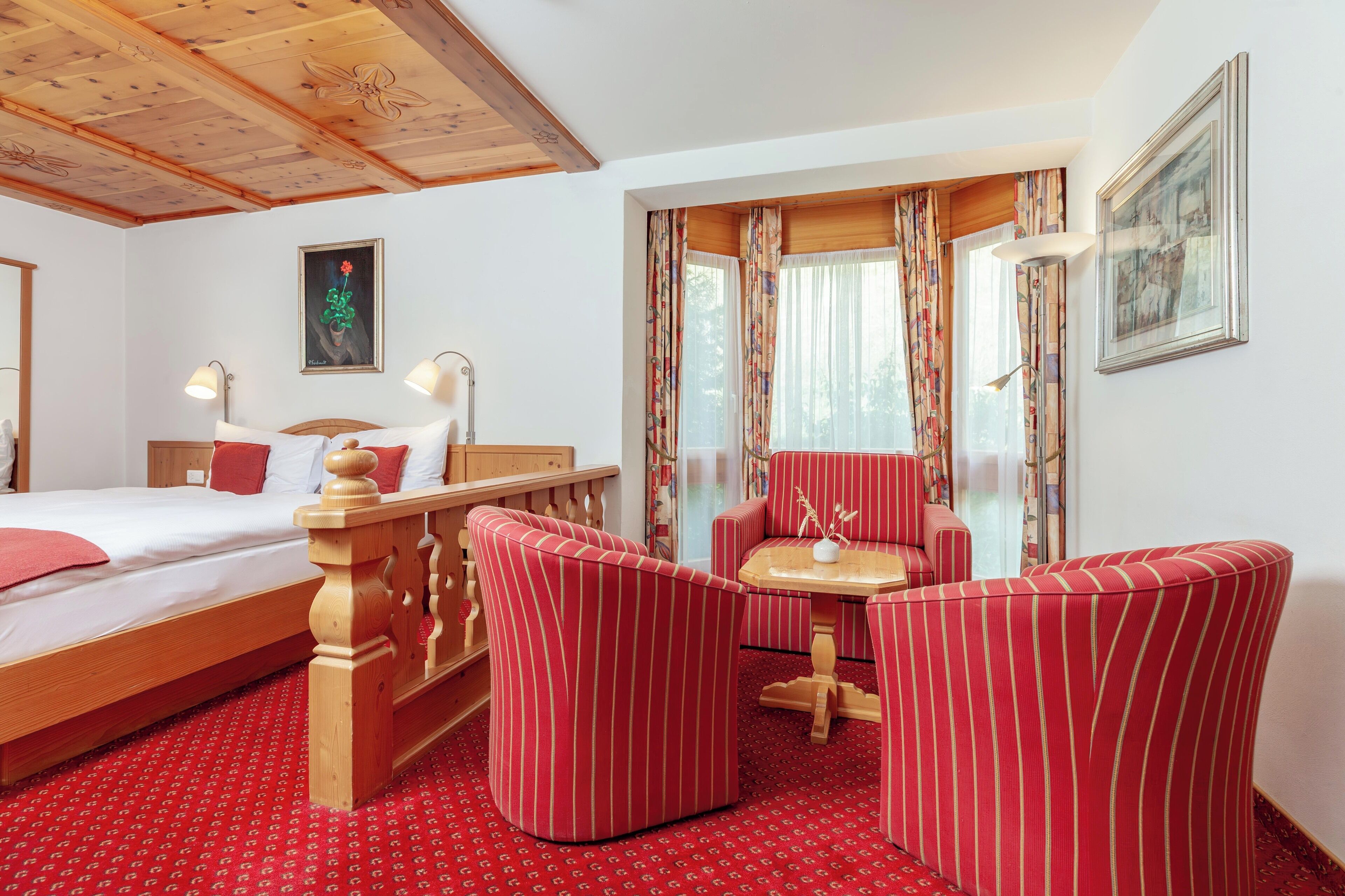 A hotel room with a wooden bed, red-striped armchairs, a small table, and a window with curtains.