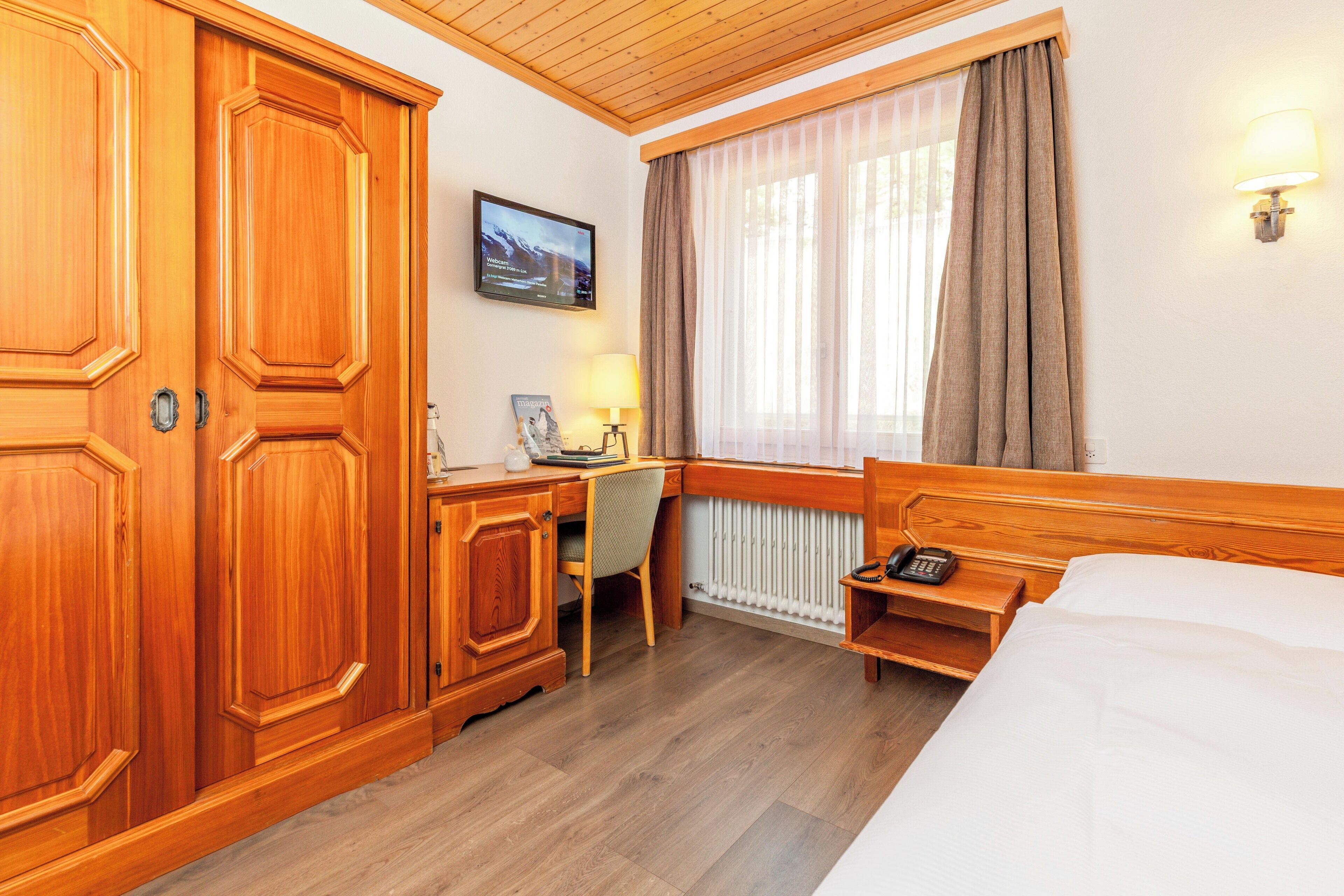 Une chambre d’hôtel meublée en bois, avec un bureau, un lit, une fenêtre avec des rideaux et un téléviseur fixé au mur.