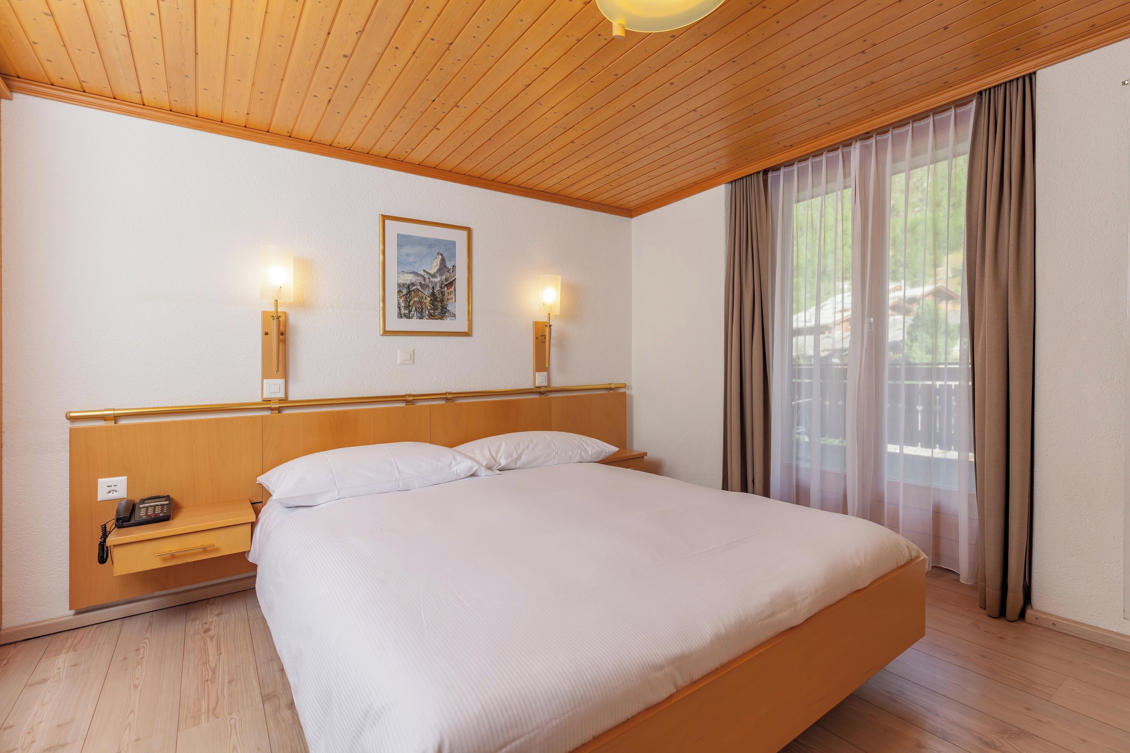 Habitación de hotel con techo de madera, una cama con ropa blanca, dos lámparas de noche, una mesita de noche con un teléfono y una ventana con cortinas.