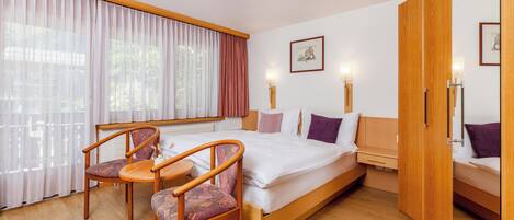 Habitación de hotel con cama, dos mesitas de noche, una silla, una mesita pequeña y una ventana con cortinas.