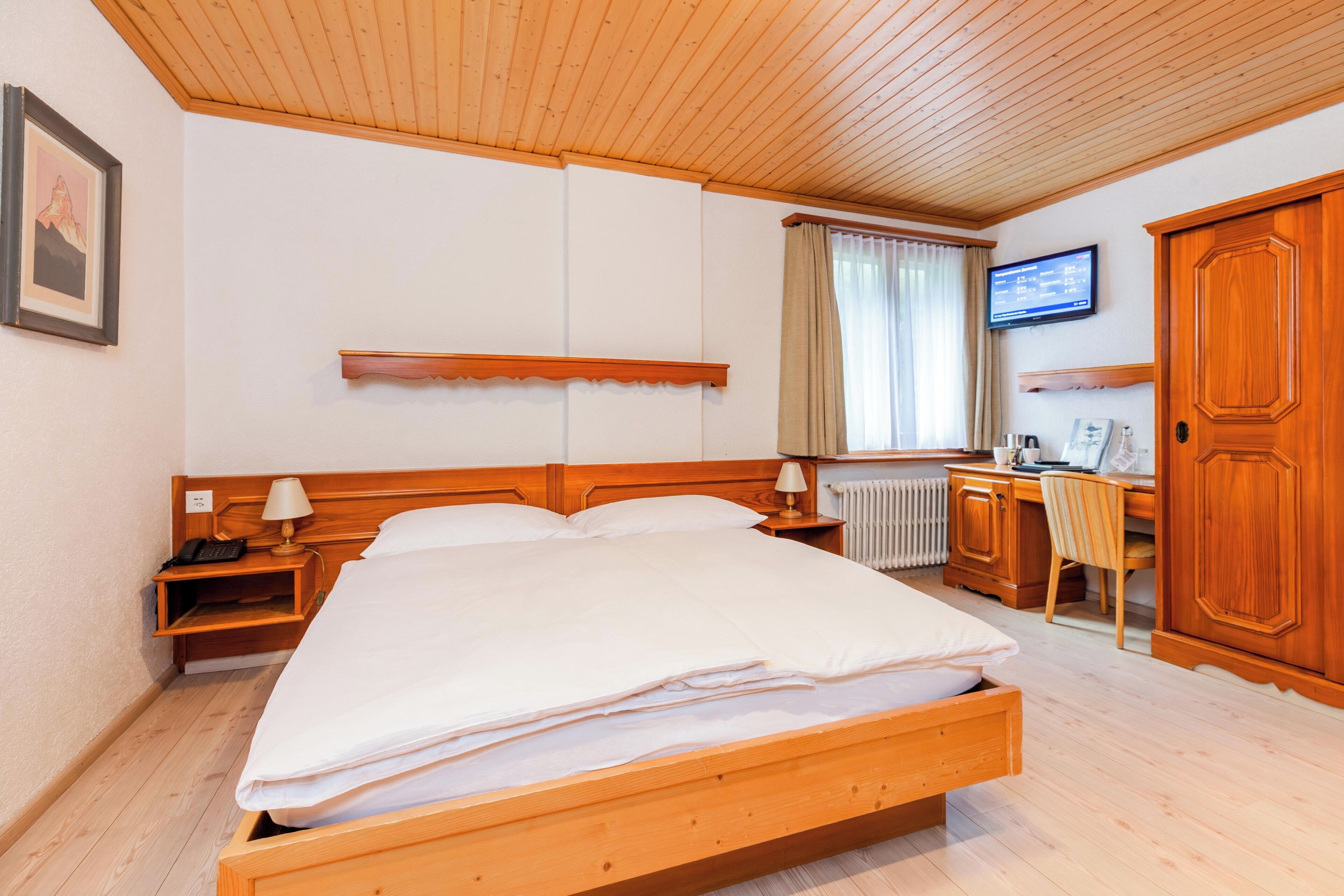 A hotel room with a wooden bed, a desk with a chair, a television on the wall, and a wardrobe.