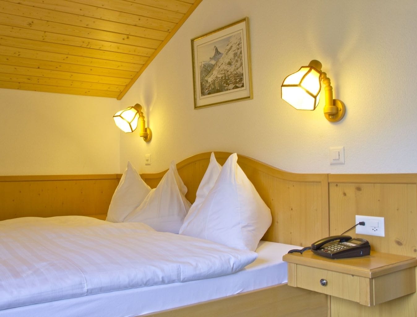 Hotellihuone, jossa on puinen katto, valkoisilla lakanoiden peitetty sänky, kaksi seinälle asennettua lamppua, sängyn vieressä oleva puhelin ja seinällä oleva kehystetty kuva.