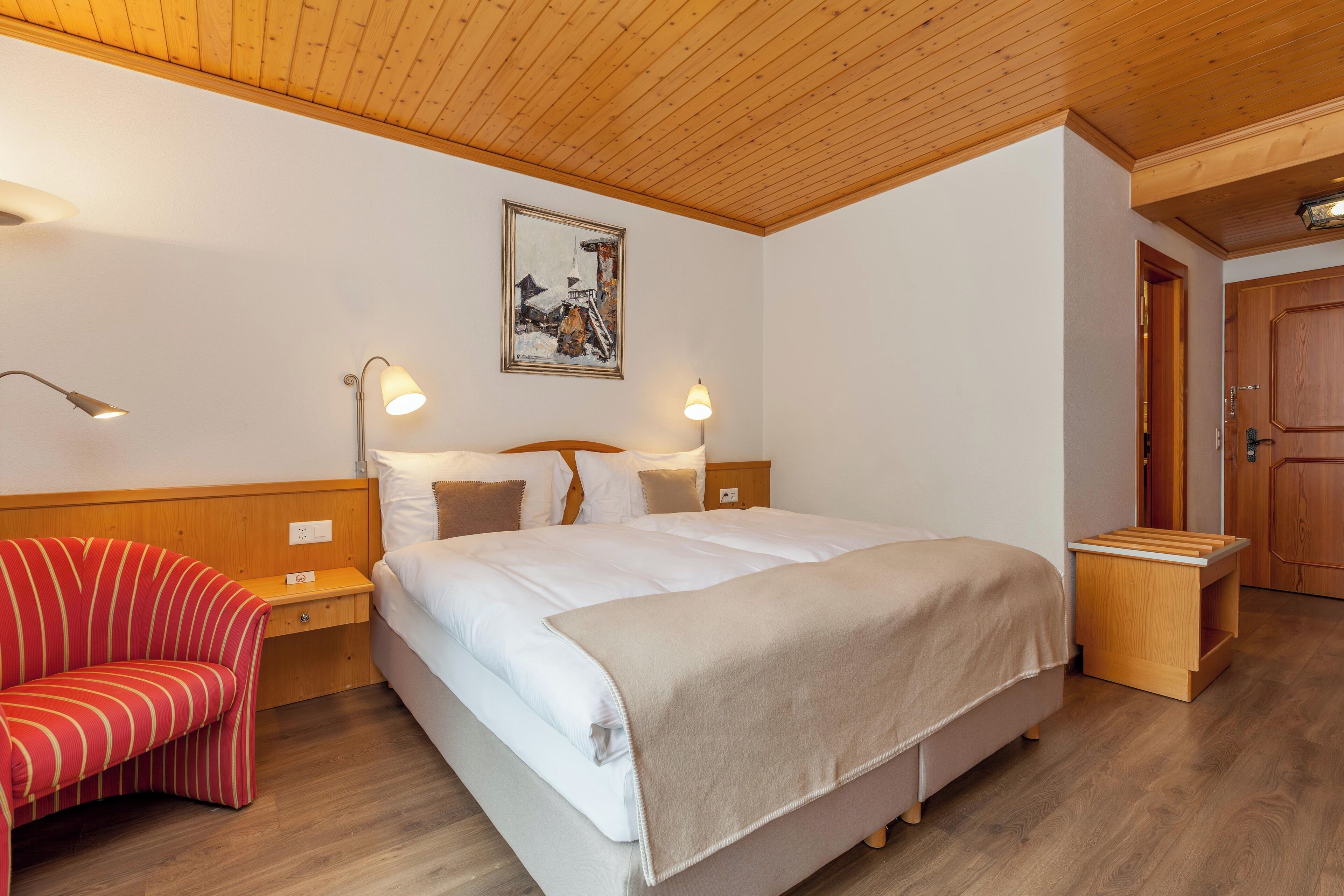 Een slaapkamer met een bed, nachtlampjes, een rode gestreepte stoel, een houten hoofdeinde en een schilderij aan de muur.
