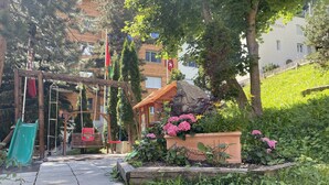 A garden area with a wooden swing set, a slide, and a planter with pink flowers. - Alpenblick (Zermatt)