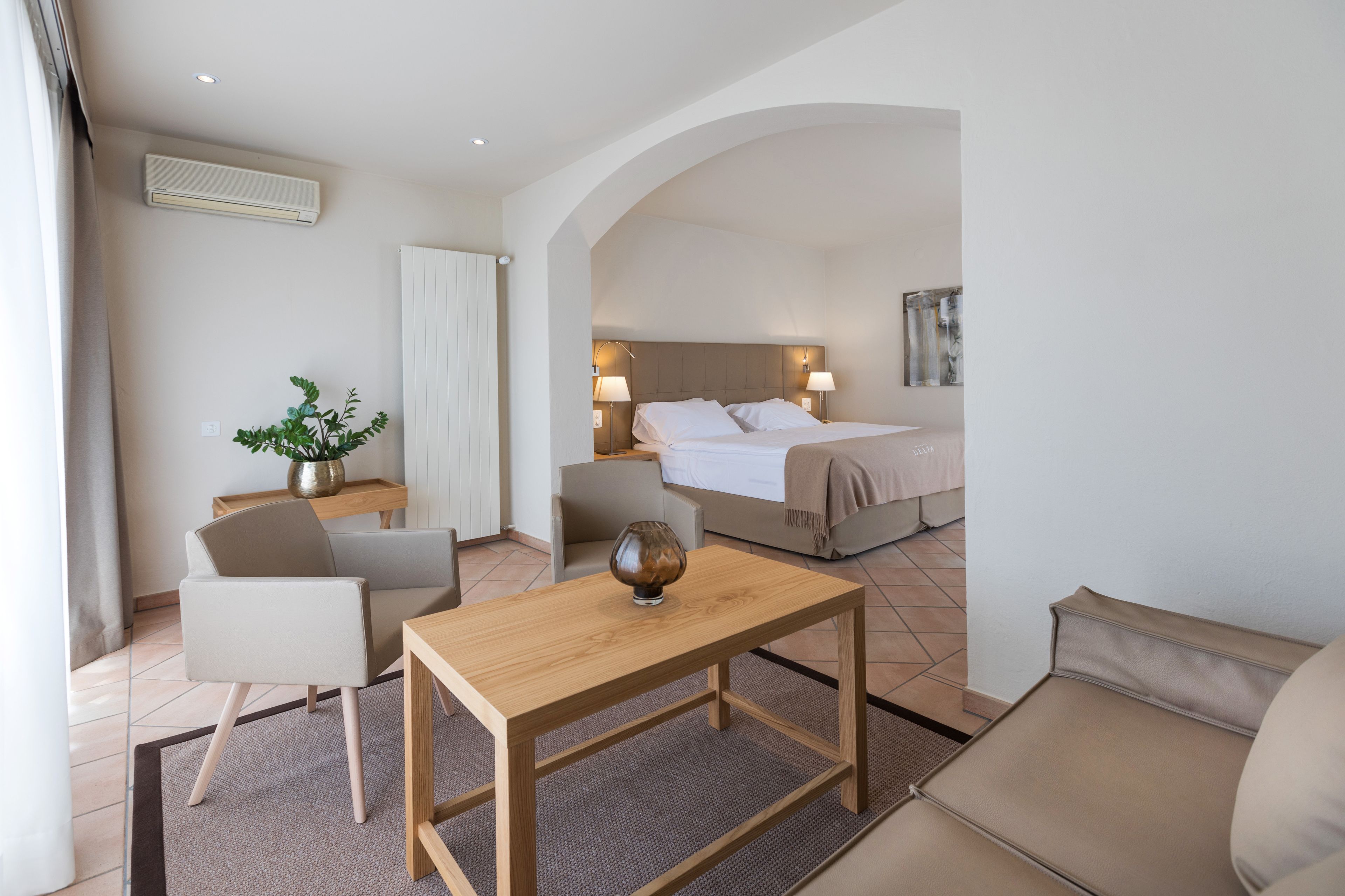 A hotel room with a bed, a sitting area with a table and chairs, a potted plant, and a large window.