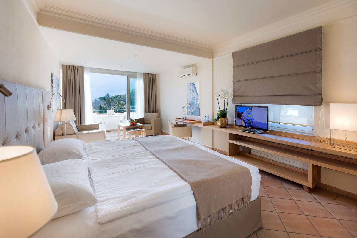 Une chambre d’hôtel avec un grand lit, un bureau avec un ordinateur, une chaise, une télévision et un balcon avec des rideaux.