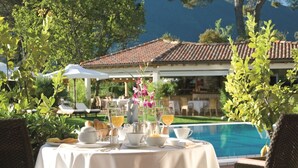 An outdoor dining area with a table set for breakfast, overlooking a swimming pool and a building with a tiled roof.