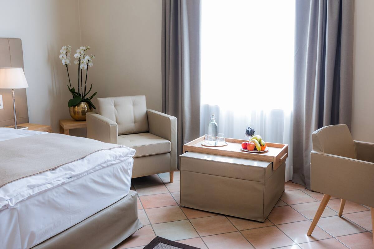 Une chambre d’hôtel avec un lit, deux fauteuils, une petite table avec des fruits, un vase de fleurs et une lampe.