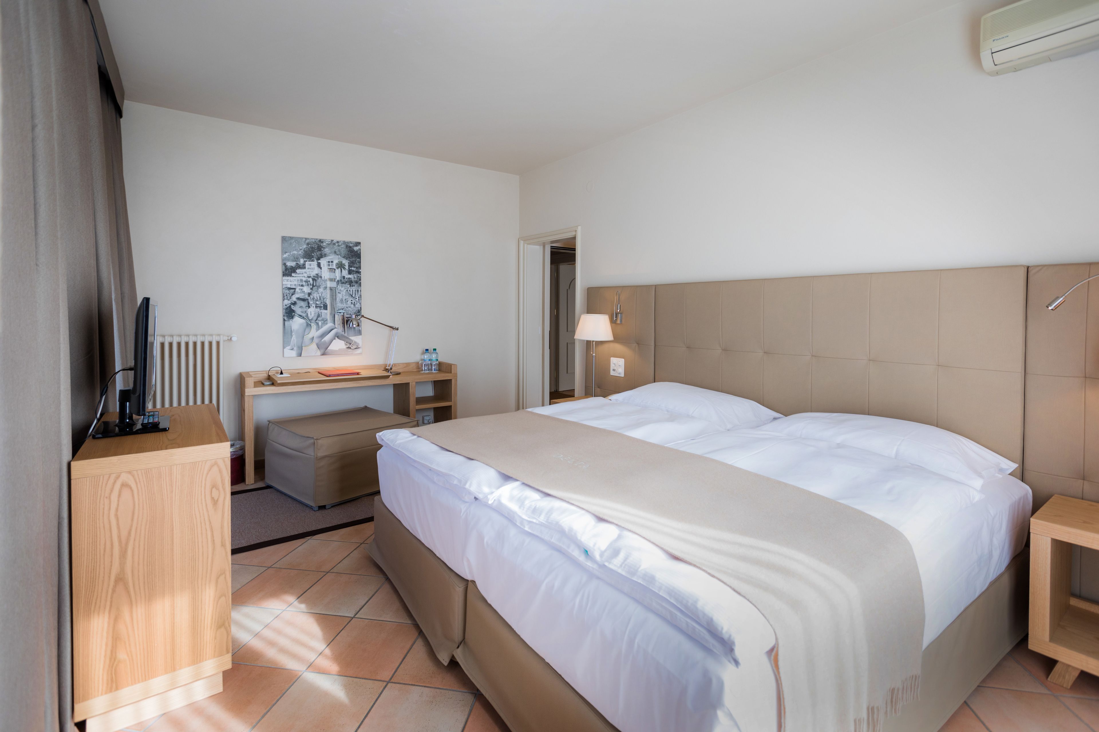 A hotel room with a bed, a desk, a chair, a lamp, and a radiator.
