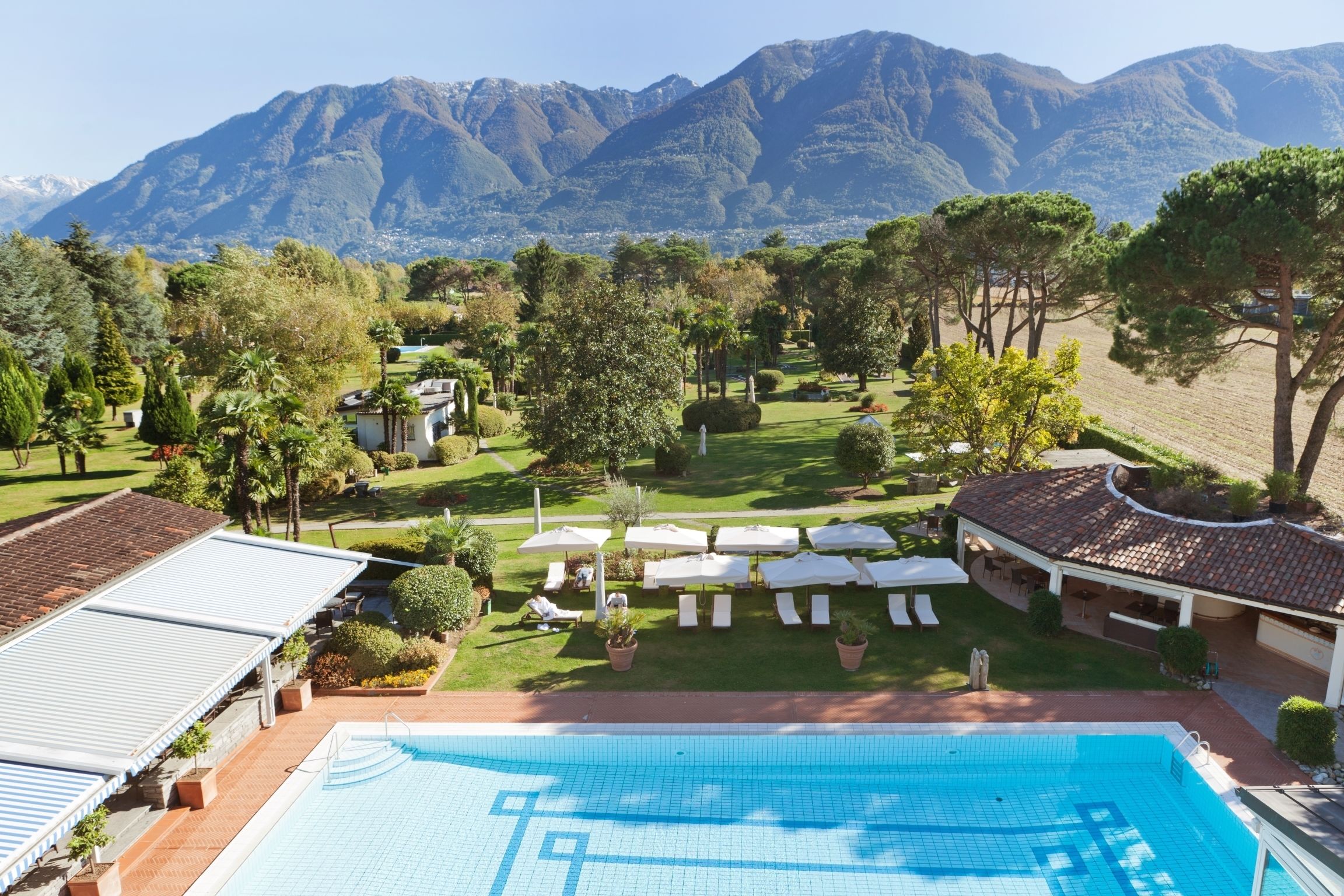 A swimming pool with a view of mountains, surrounded by a garden with lounge chairs and umbrellas.