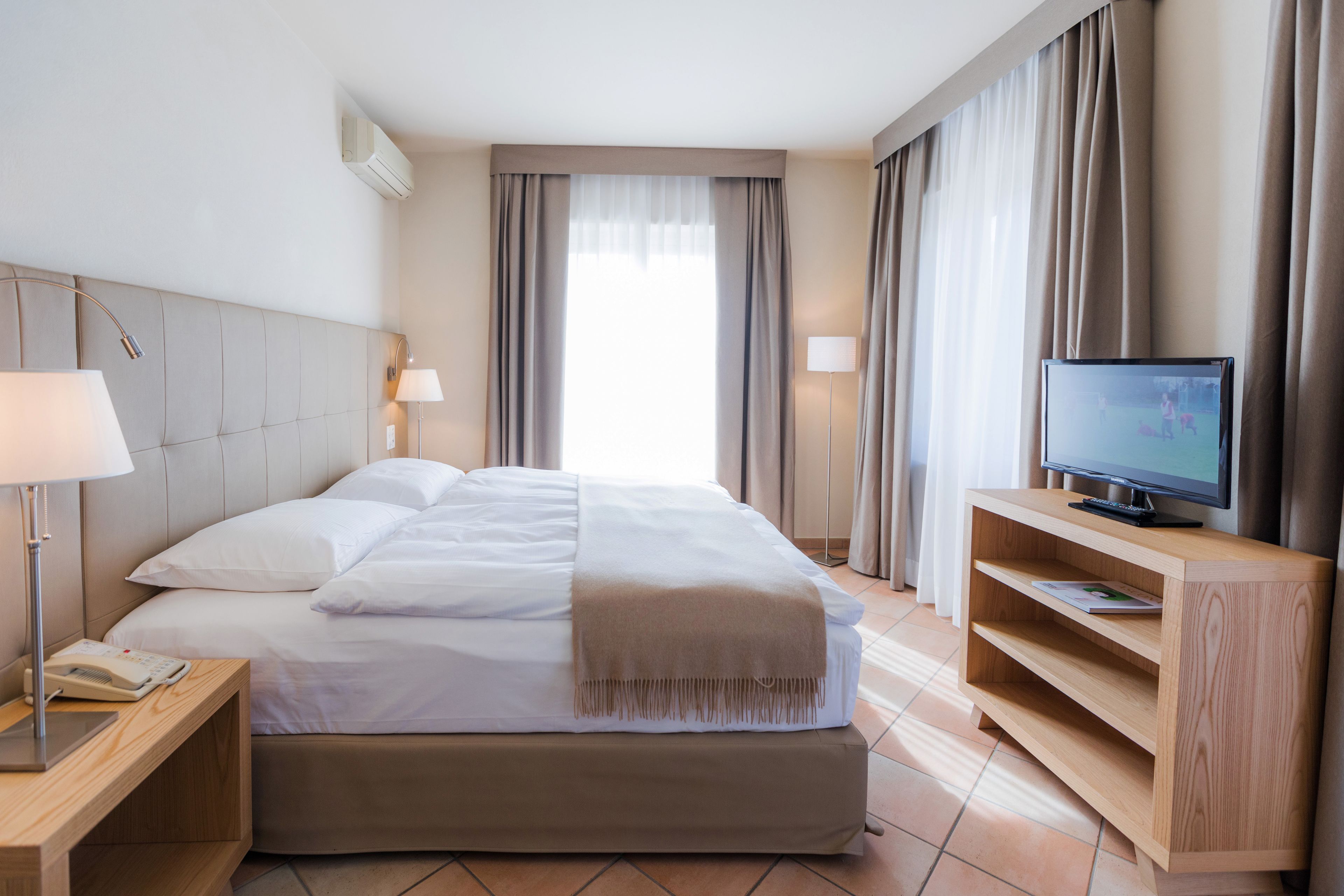 A hotel room with a bed, bedside lamps, a TV on a wooden stand, and a window with curtains.
