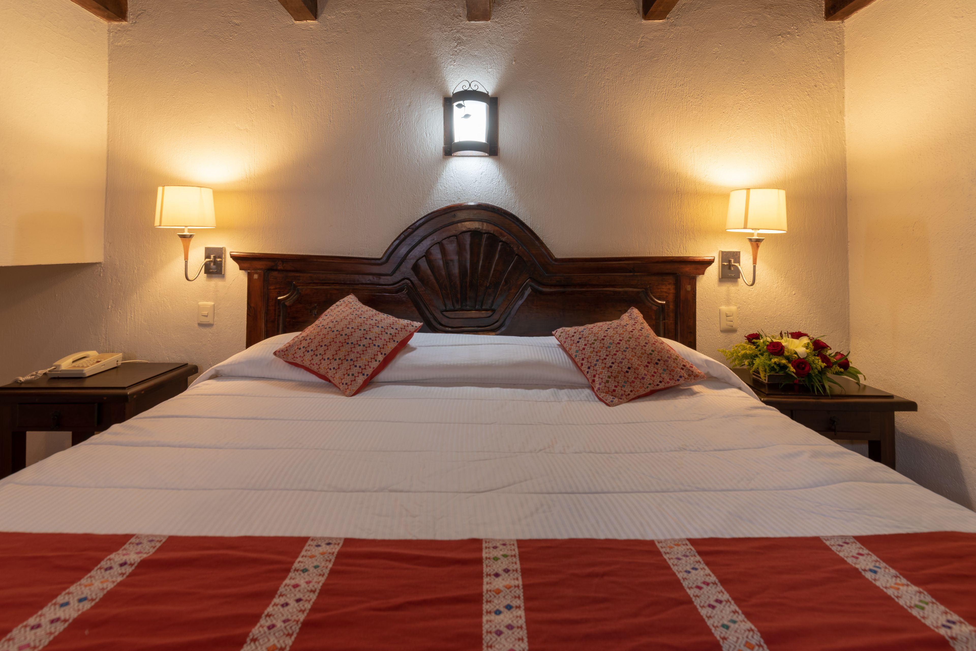 A neatly made bed with a wooden headboard, two bedside lamps, a nightstand with a phone, and a vase with flowers on the right side.