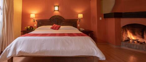 A bedroom with a large bed, two bedside lamps, a nightstand with a phone, a fireplace, and a wall clock.
