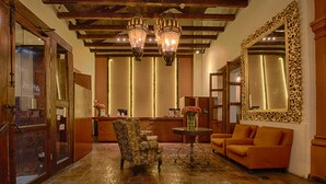 A lobby with a wooden reception desk, a patterned armchair, a round table with a vase, and a large ornate mirror. - Hotel Plaza Magnolias (San Cristóbal de las Casas)