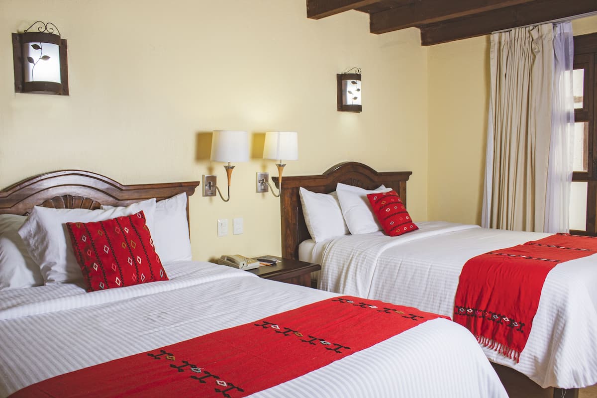 Une chambre d’hôtel avec deux lits, chacun agrémenté de draps blancs et de coussins décoratifs rouges. On y trouve des lampes fixées au mur et une petite table de chevet avec un téléphone.
