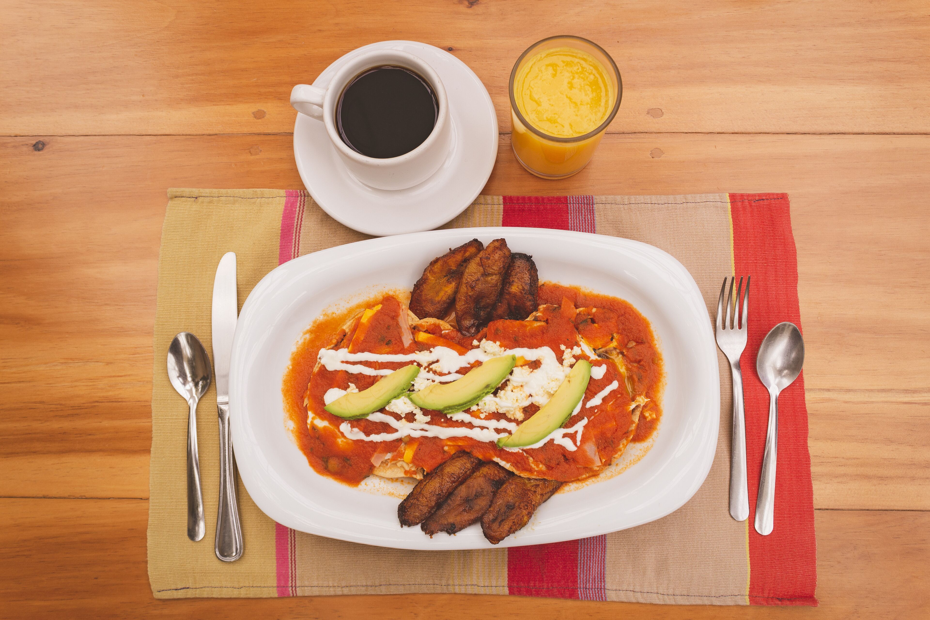 A plate of enchiladas with avocado slices and a side of black coffee and orange juice on a wooden table.