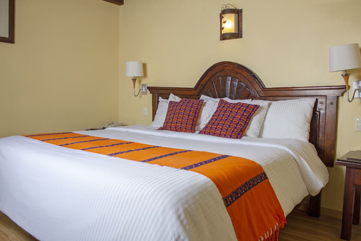 Un lit bien fait, recouvert d’une couvre-lit blanche et orange, agrémenté de deux coussins à motifs et doté d’une tête de lit en bois.