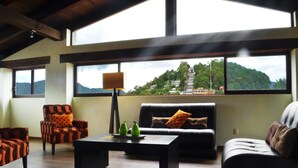 A modern living room with a sofa, armchairs, and a coffee table. Large windows offer a view of a mountain and a pagoda. - Hotel Plaza Magnolias (San Cristóbal de las Casas)
