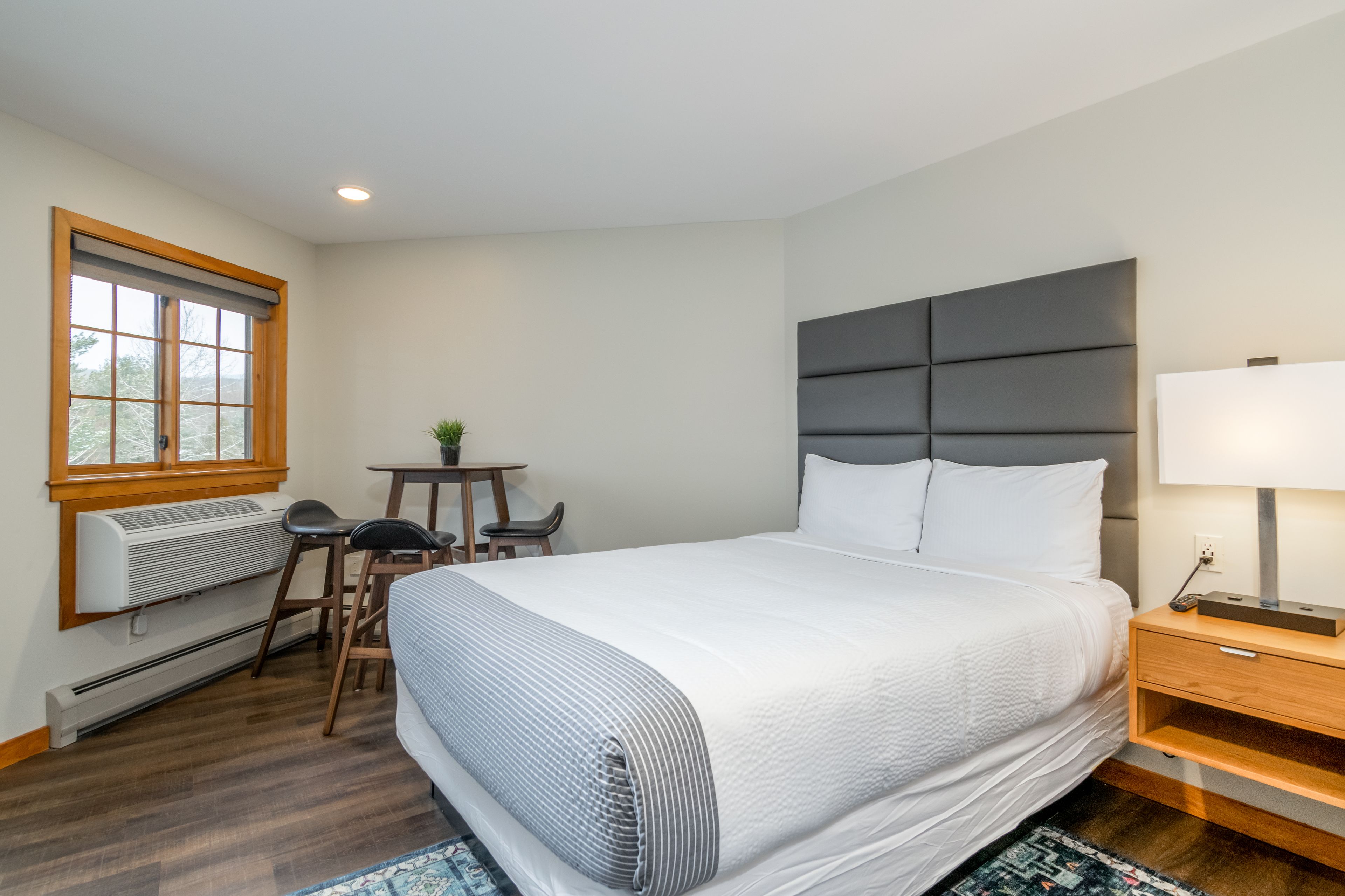 A neatly arranged hotel room with a large bed, a bedside table with a lamp, a small table with chairs, and a window with blinds.