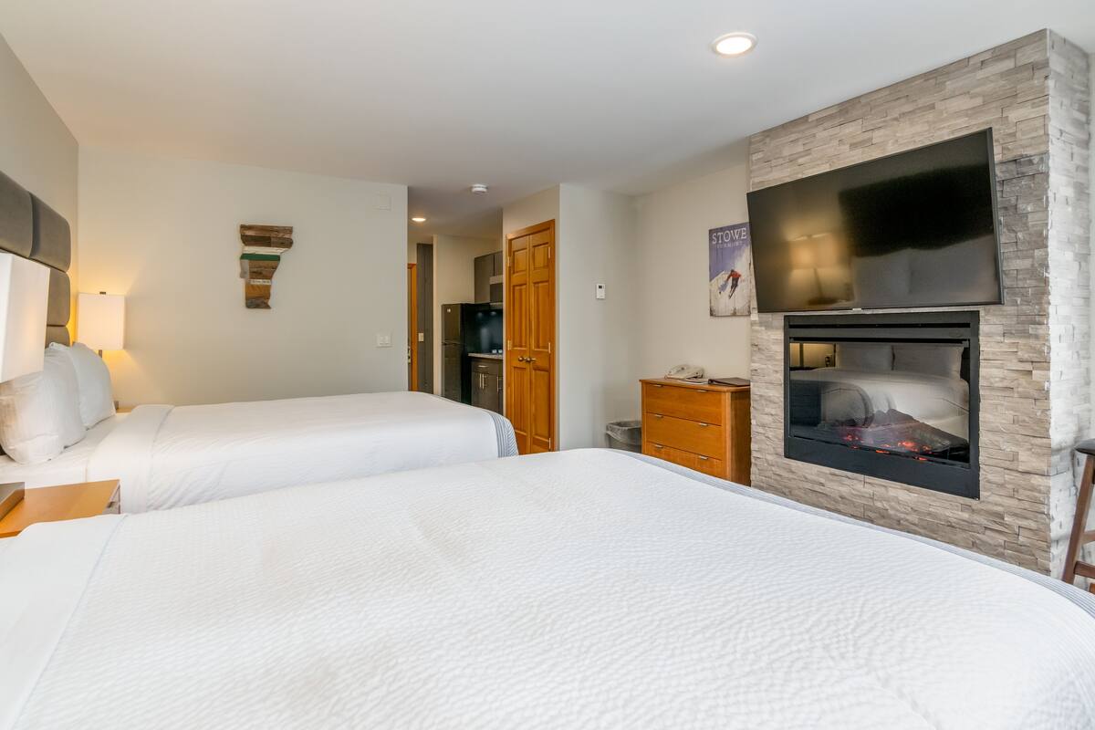 A hotel room with two beds, a TV mounted on the wall, a fireplace, and a wooden wardrobe.