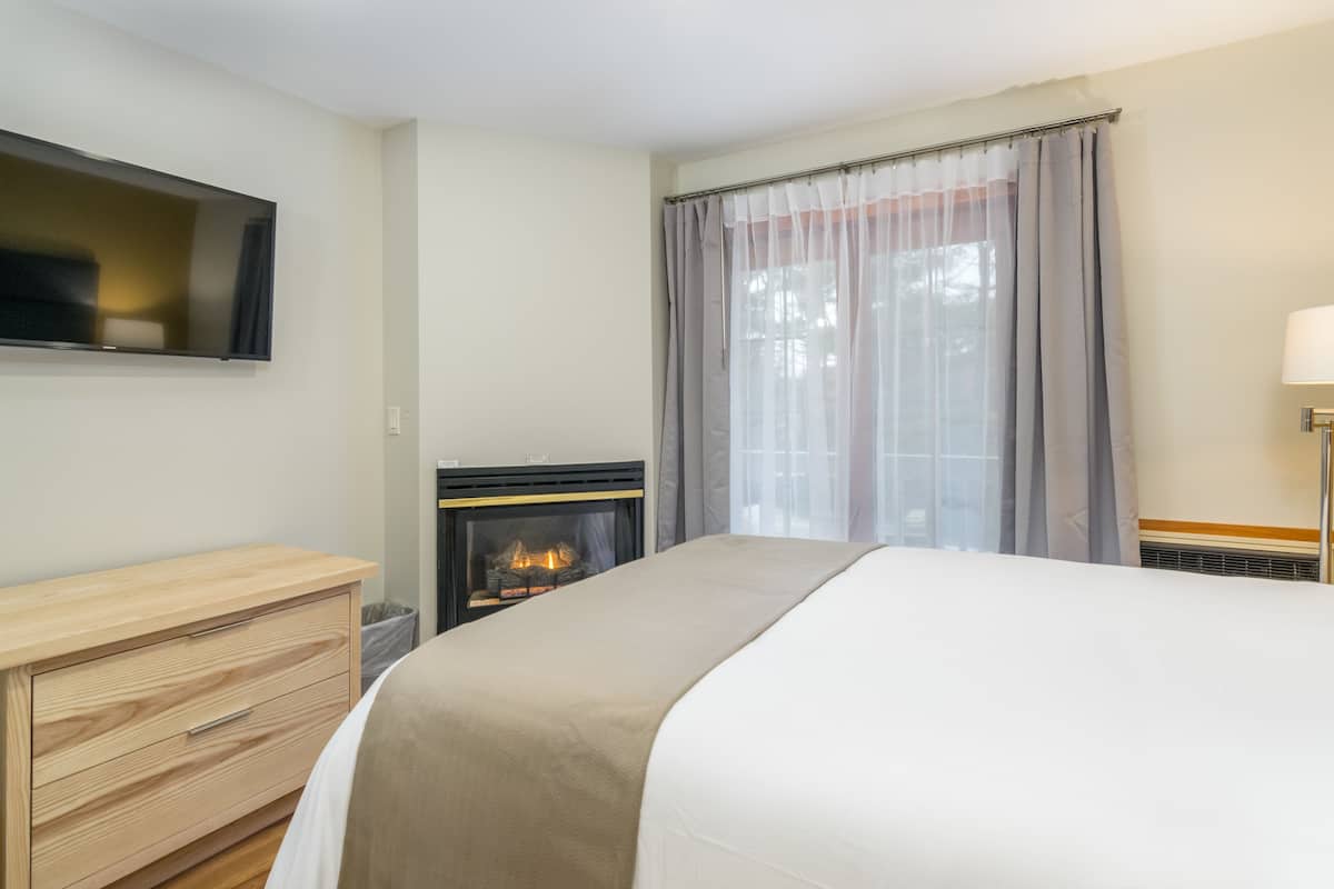 A bedroom with a bed, a wooden dresser, a fireplace, a television mounted on the wall, and a window with curtains.