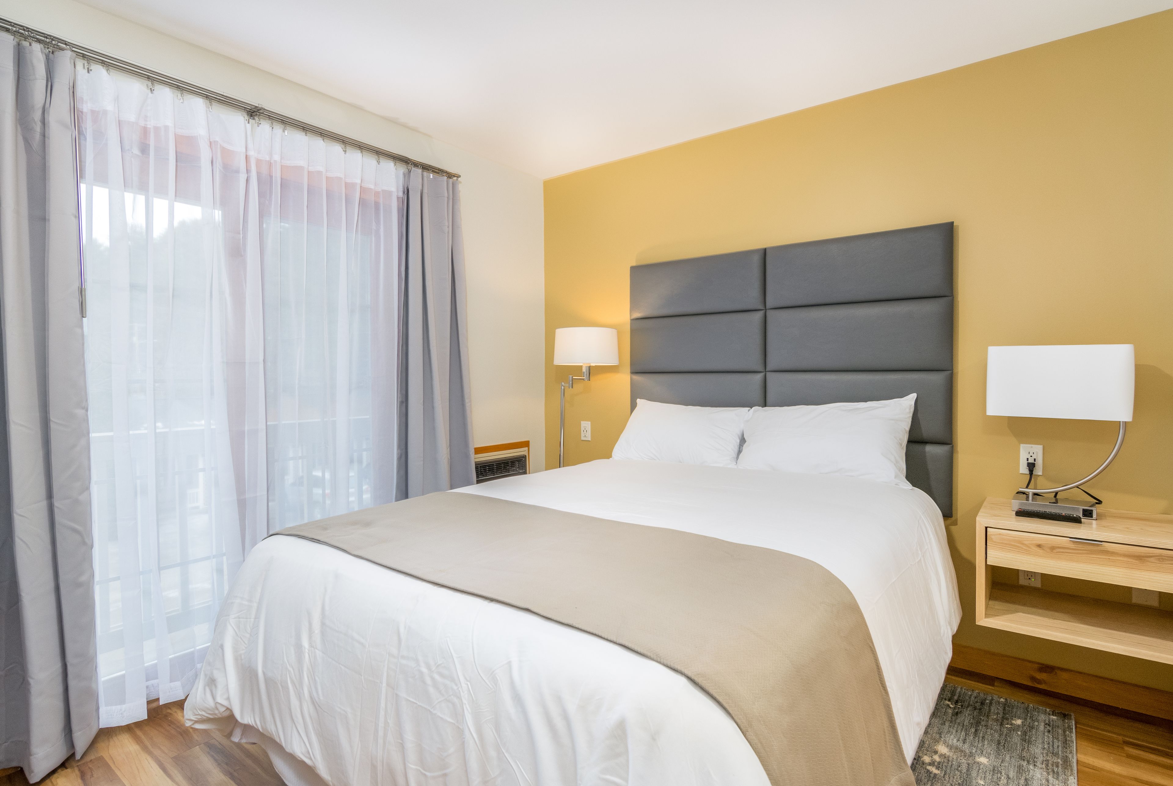 A hotel room with a large bed, a bedside table with a lamp, and a window with sheer curtains.