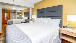 A hotel room with two beds, a wooden floor, a grey headboard, a bedside table with a lamp, and a wall-mounted TV.