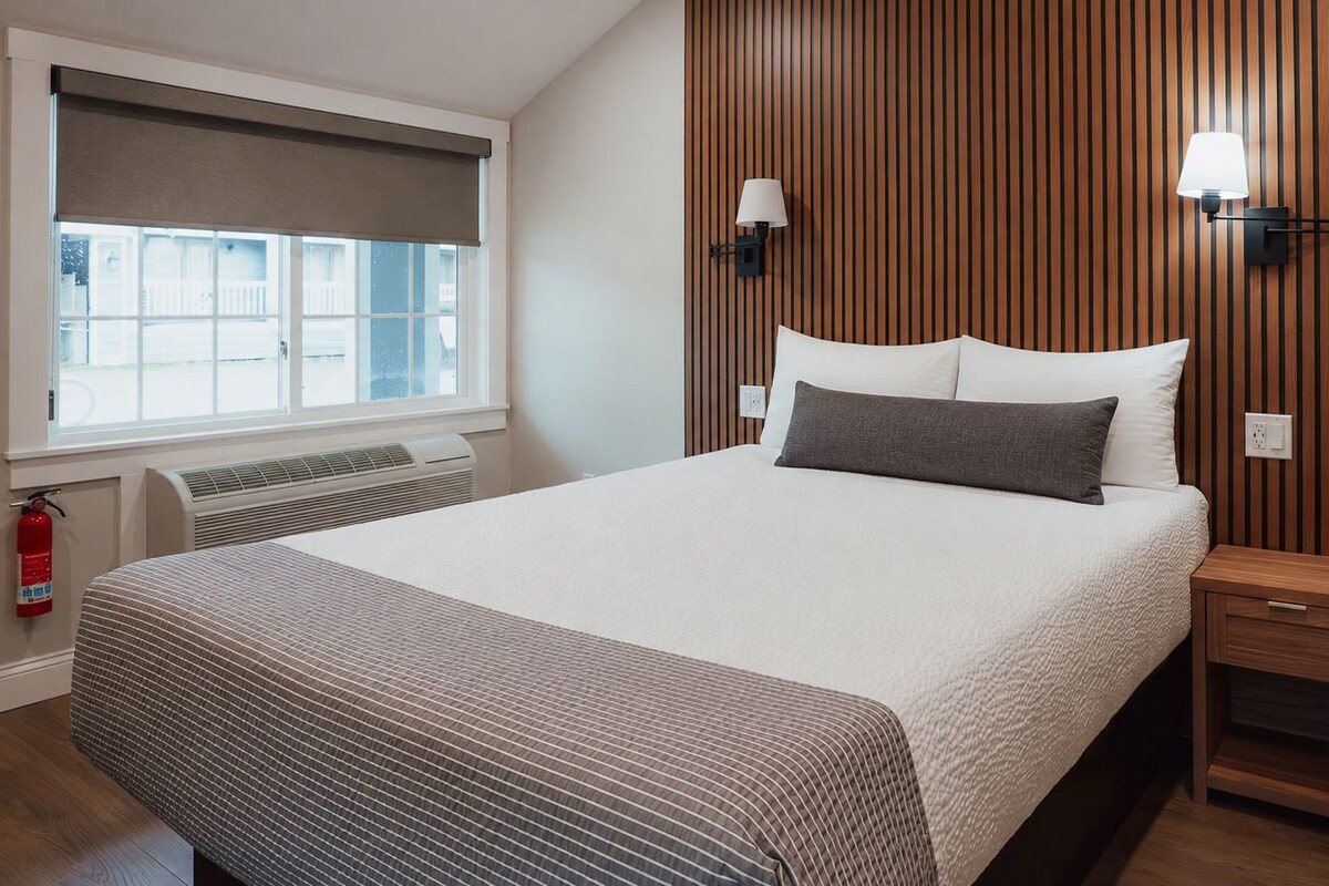 Una habitación de hotel con cama, mesitas de noche, ventana con persianas, lámpara de pared y extintor.