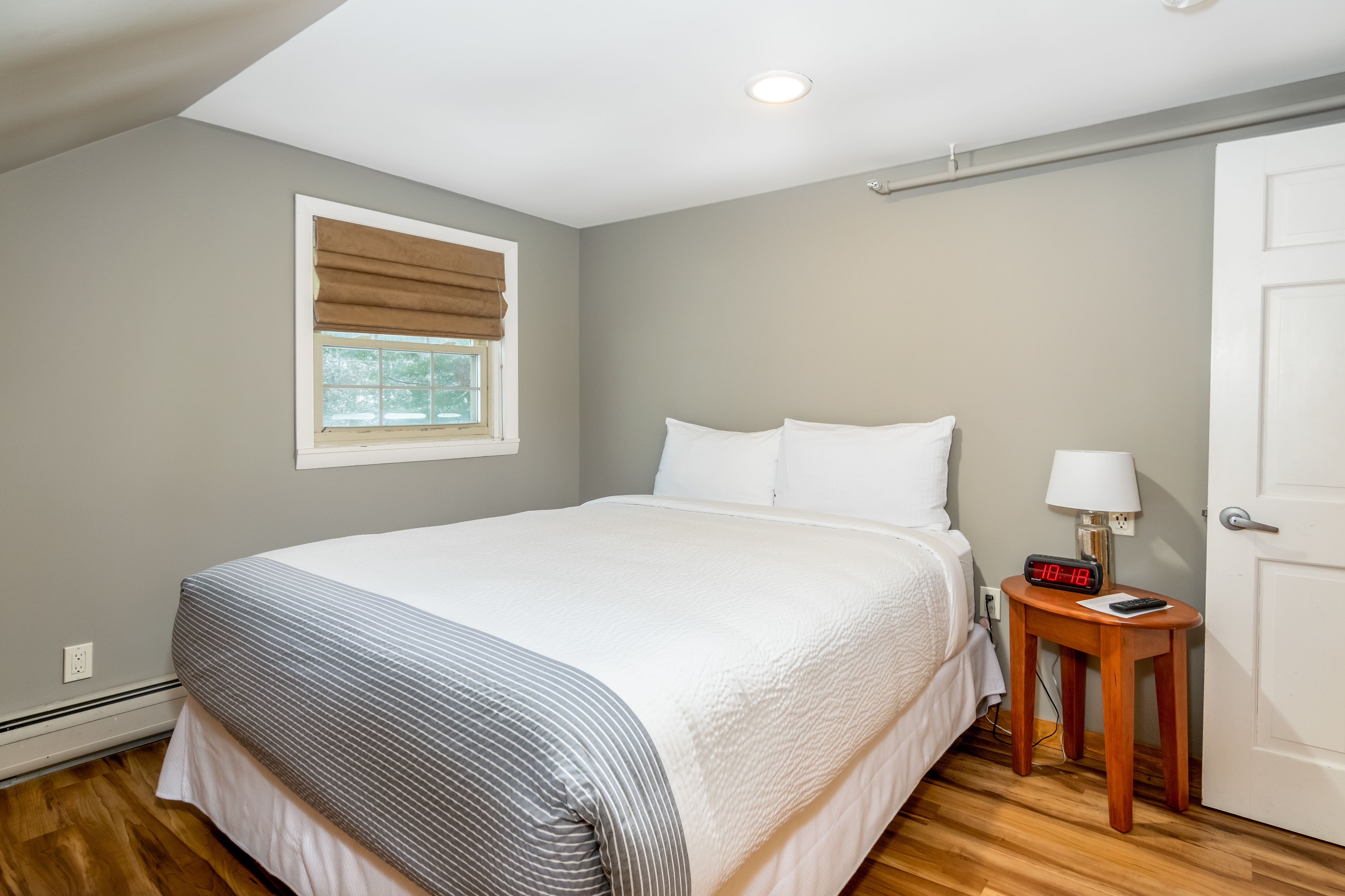 A bedroom with a bed, a bedside table with a lamp, a clock, and a window with blinds.