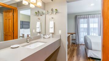 A modern bathroom with a large mirror, two sinks, and a shower. There is a bed visible in the adjacent room.