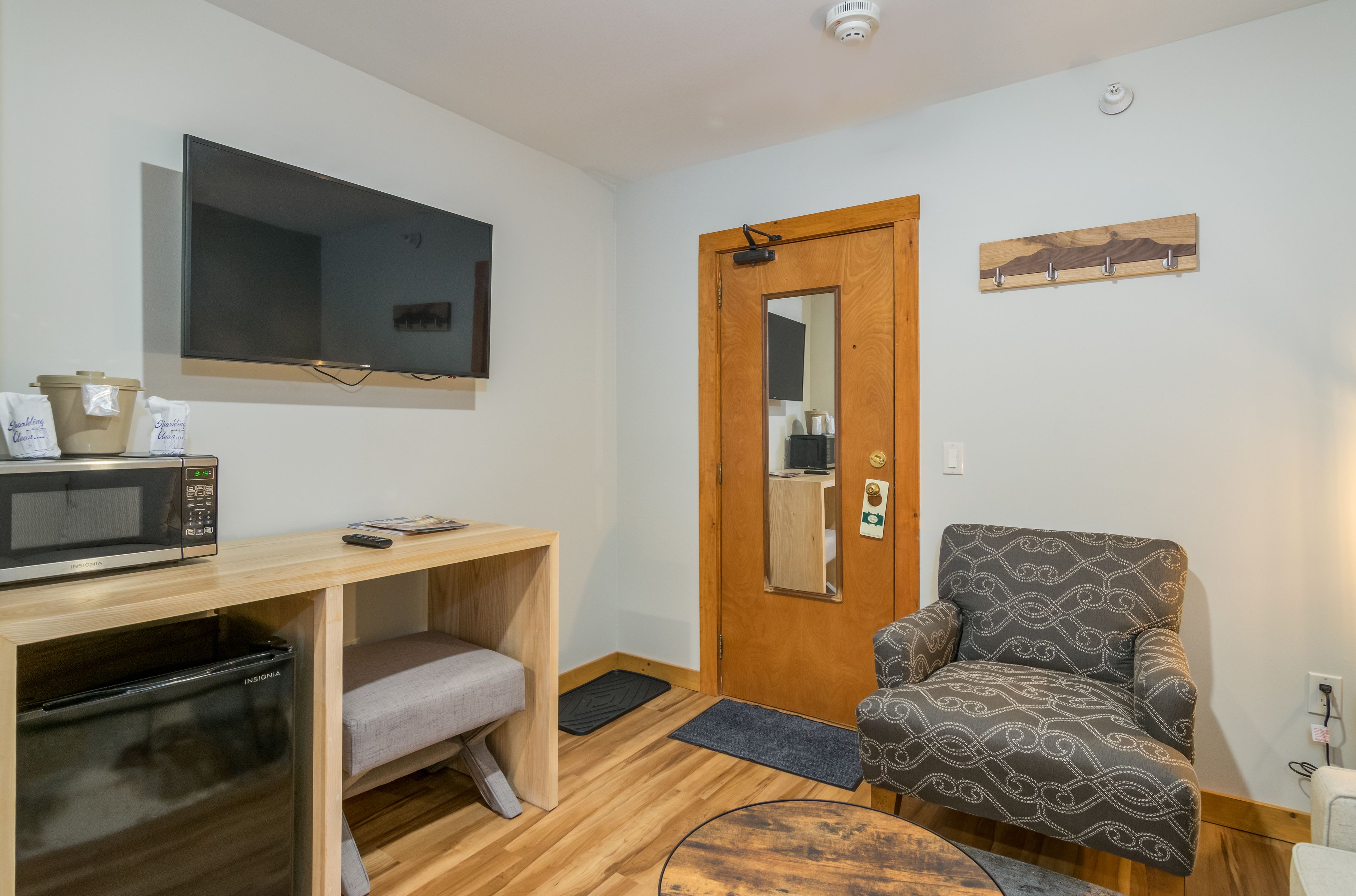 A compact room with a wooden desk, a microwave, a refrigerator, a gray chair, a wooden door, and a mounted television.
