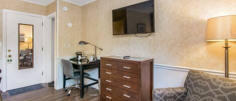 A room with a desk, chair, and a sofa. There is a TV mounted on the wall and a lamp on the floor.