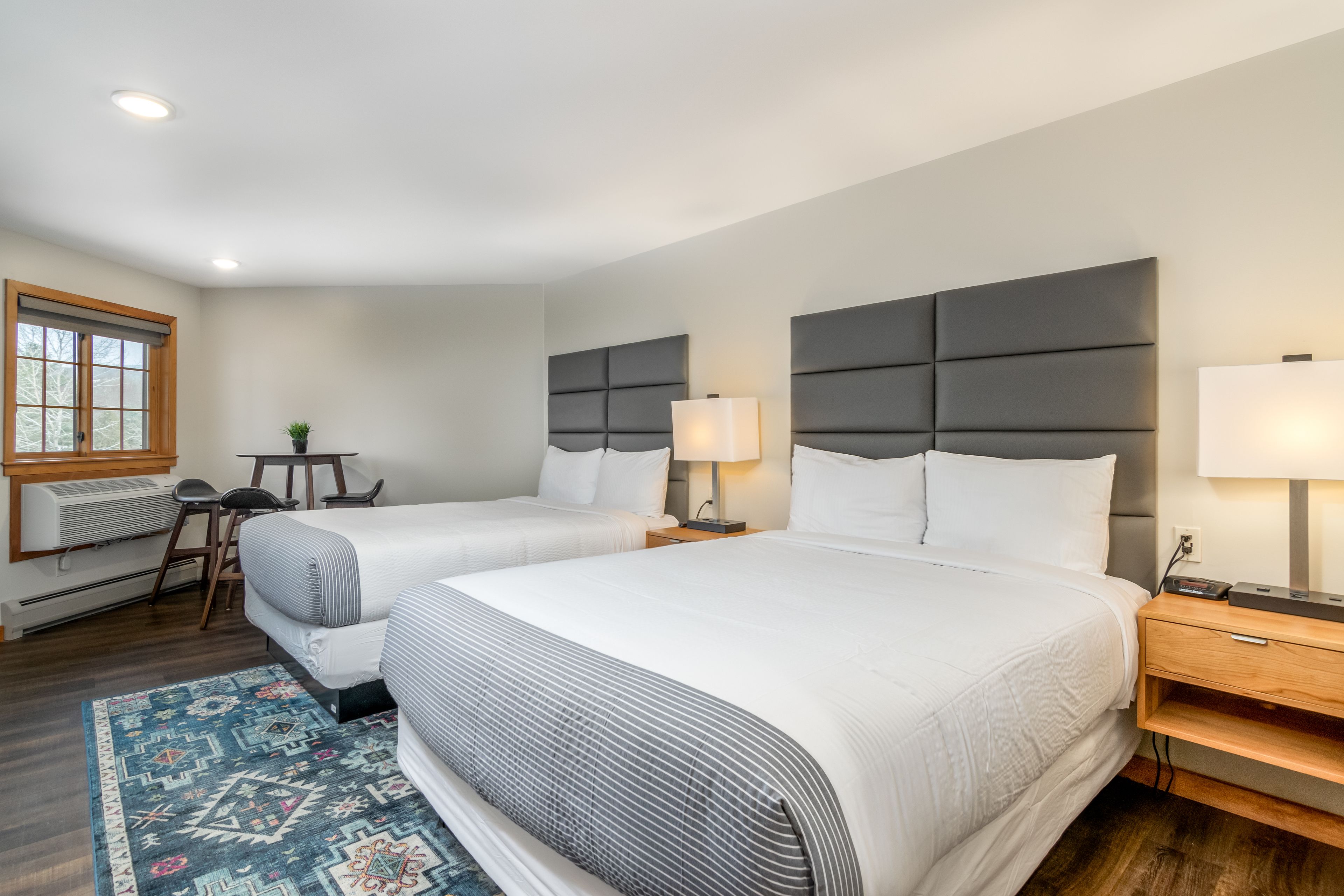 A hotel room with two beds, a desk, a chair, a small table, and a window with blinds.