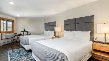 A hotel room with two beds, a desk, a chair, a small table, and a window with blinds.