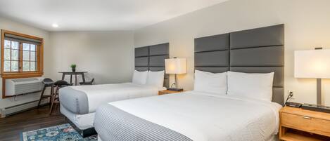 A hotel room with two beds, a desk, a chair, a small table, and a window with blinds.