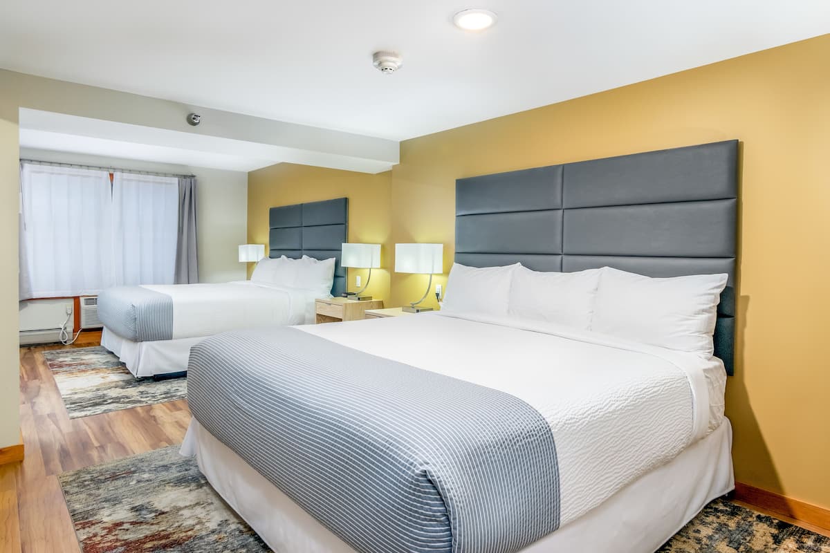 A hotel room with two beds, a large window with curtains, a bedside table with lamps, and a headboard.