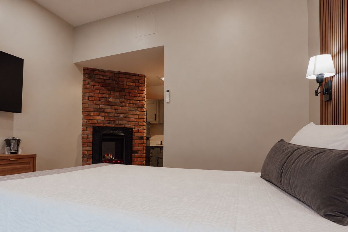 Habitación de hotel con una cama grande, una chimenea de ladrillo, un televisor de pantalla plana montado en la pared y una lámpara de noche.