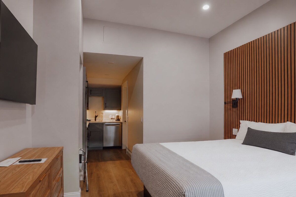 Habitación de hotel moderna con cama, mesita de noche, televisión y vistas al área de la cocina.