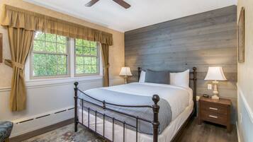 Un dormitorio con cama, dos mesitas de noche, ventilador de techo, ventana con cortinas y vista a follaje exterior.