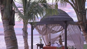 Un mirador con dosel, una cama y una mesita de noche con lámpara, frente a un telón de fondo de playa.